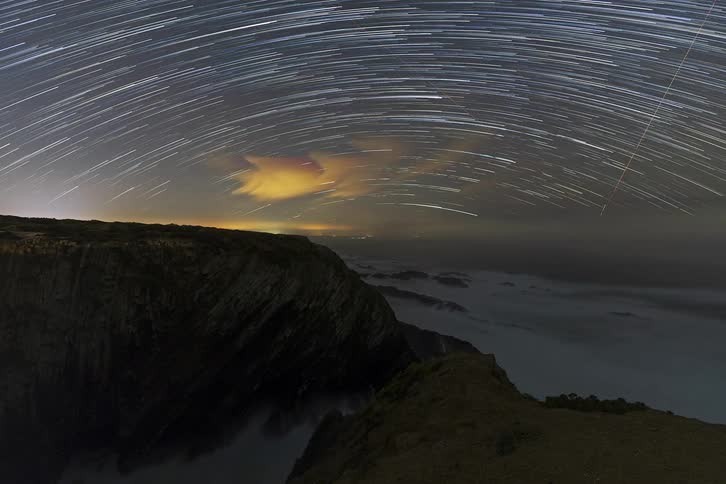 Long exposure star trails arc over a dark rocky coastline where thick mist settles in the cliffs and waves crash against the shore under a deep night sky featuring distant orange glows on the horizon