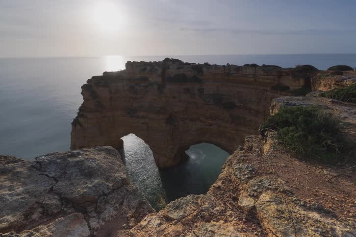 The video showcases a wide shot of a rugged coastal cliff featuring a natural stone double arch overlooking the ocean as the sun shines brightly in the sky a person can be seen walking along the top edge of the rocky formation