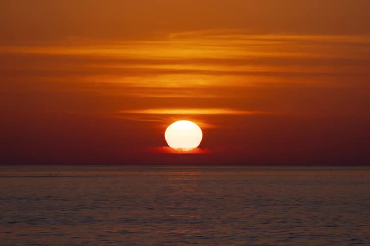Sun setting on the horizon over the sea under an orange sky with layered clouds