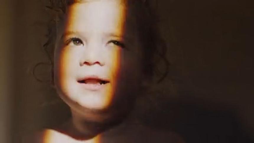 A curly haired toddler smiles in a dark room while a warm light glow illuminates the side of her face and hair as she looks toward the side of the frame in a close up shot