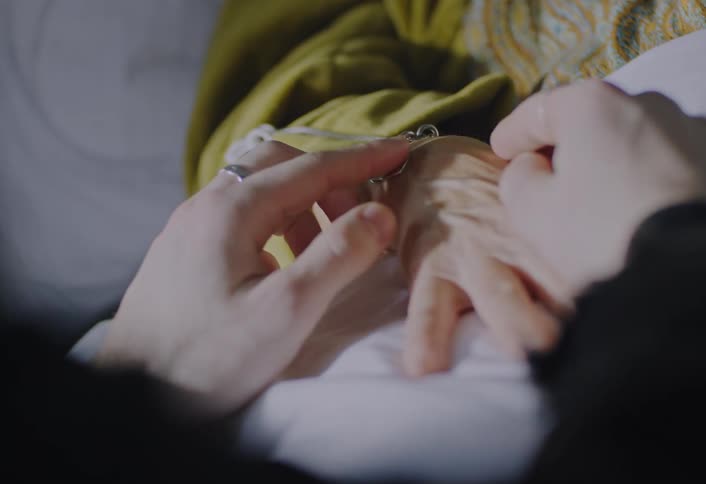 A close view shows a younger person gently caressing the wrist of an elderly person and then holding their hand