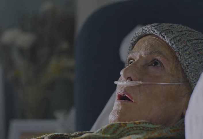 A young man sits in grief near a hospital bed where an elderly woman with an oxygen tube is resting