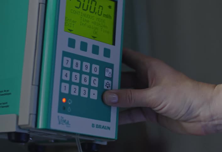 A woman with a wedding ring and silver bracelet uses her hand to press buttons on a b braun medical infusion pump in a dimly lit clinical setting