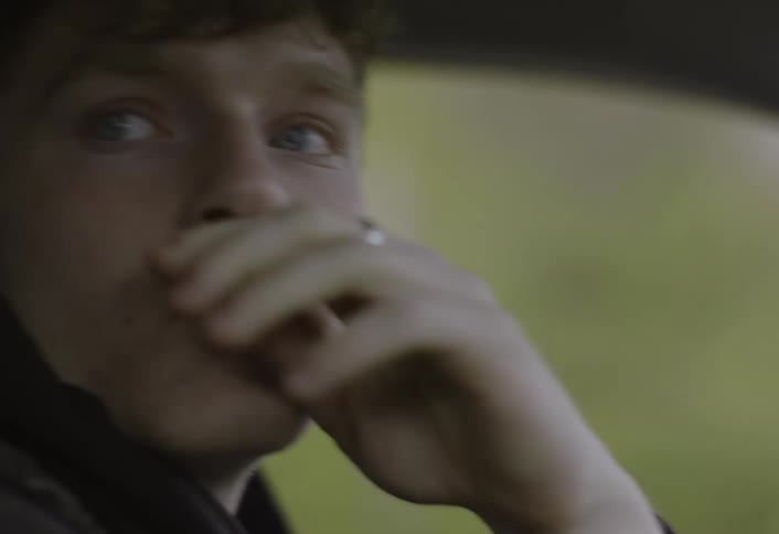 A young man with short brown hair sits in the driver seat of a car and looks out the side window before turning his head to look forward as green trees pass by in the blurred background