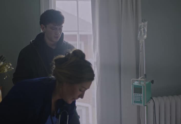 In the quiet hospital room a nurse leans over a patients bed while talking with a young man standing nearby the soft daylight from the window illuminating the iv drip beside them