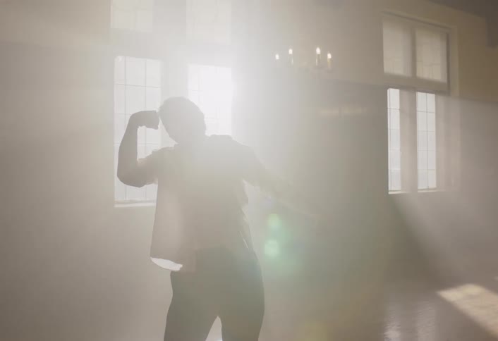 Woman stands in a large bedroom watching a young man dance as bright sunlight shines into the room