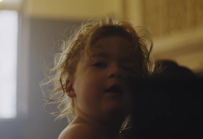 A curly haired toddler boy in a diaper climbs up to a black upright piano to touch the keys and a red rose while framed photos sit on top of the instrument in a dimly lit room