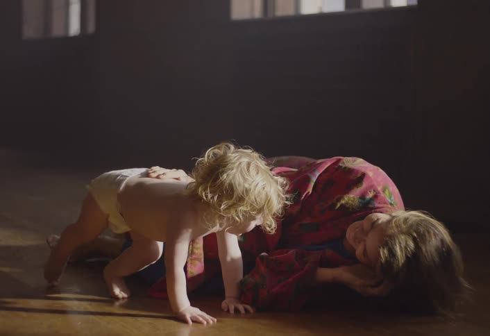 A young woman is crawling on the floor with a baby both laughing and smiling in a dimly sunlit large room