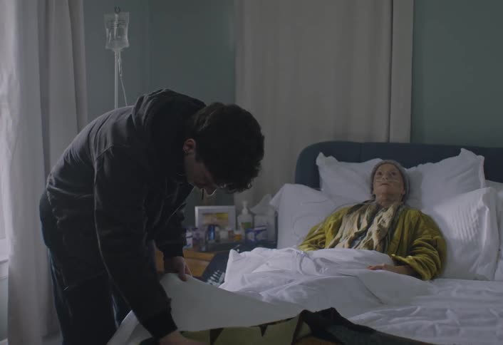 A young man stands beside the hospital bed arranging the blankets toward an elderly woman lying beneath the blankets as soft daylight filters through the window behind them