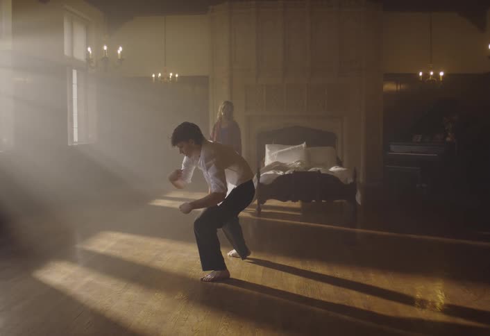 A young man in a white shirt and dark pants performs a contemporary dance in a large sunlit bedroom while a woman in a robe watches from the background near a bed and a piano