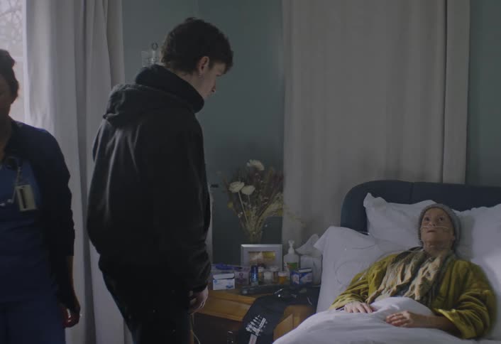 A young man in a black jacket enters a room and kneels beside an elderly woman lying in a bed who has medical tubes in her nose