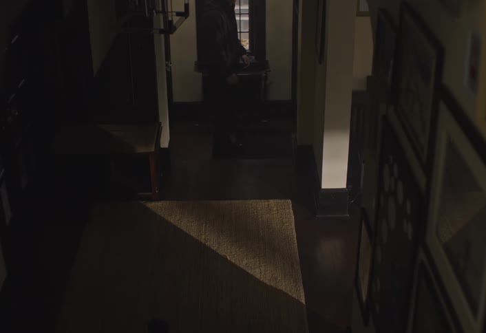 A person dressed in dark clothing walks in a dimly lit hallway of a house looking to the side as warm light spills from rooms further inside