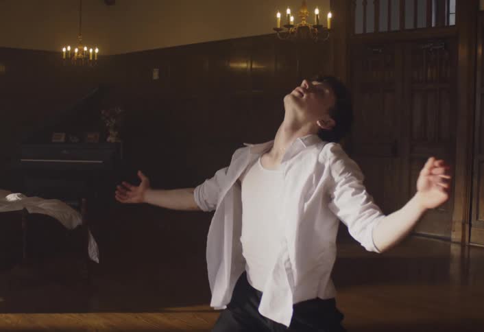 Man swings his arms as he dances and a woman sits in bed watching as sunlight shines brightly through the windows