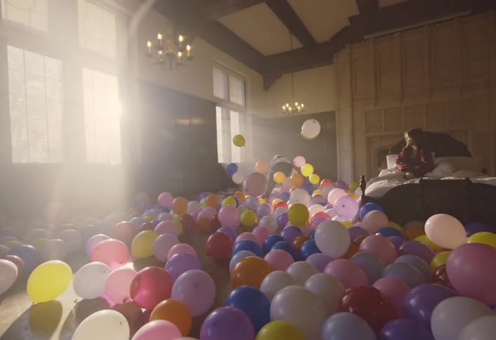 Woman sitting up in bed watches a man dance among the balloons covering the floor of the bedroom on a sunny day