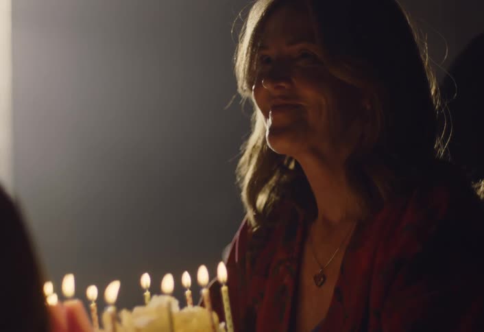In the dim smoky light a woman sits with her head bowed blowing out birthday cake candles while a shadowed figure stands before her