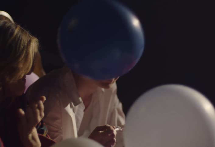 A man and a woman sit on a bed in a dark room while eating cake and laughing among many colorful balloons