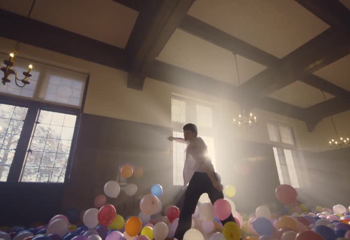 A young man dancing lively in a dimly sunlit room with a floor full of air filled colorful balloons