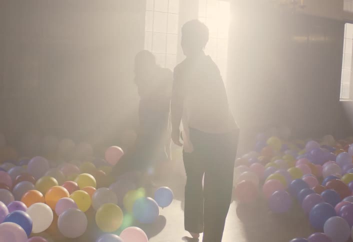 A young couple indoors in a hall filled with balloons on the floor as they dance around holding hands