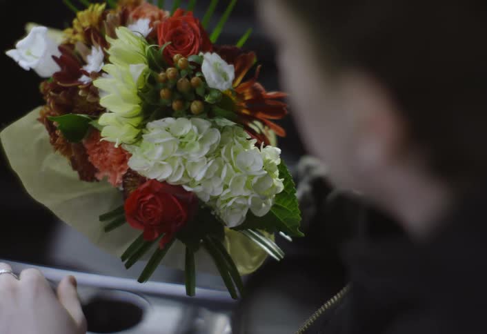 A close view shows a man sitting in the front seat of a car while he reaches for a colorful bouquet of flowers on the passenger seat