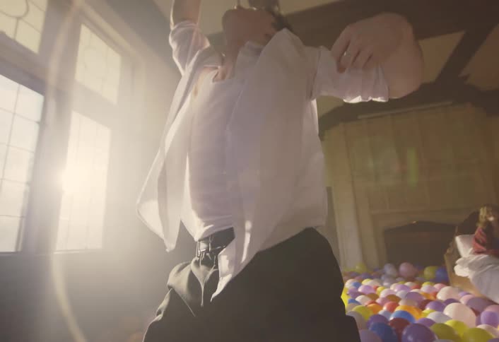 Balloons cover the floor as a young man dances and a woman sits in bed watching as sunlight shines brightly through the windows