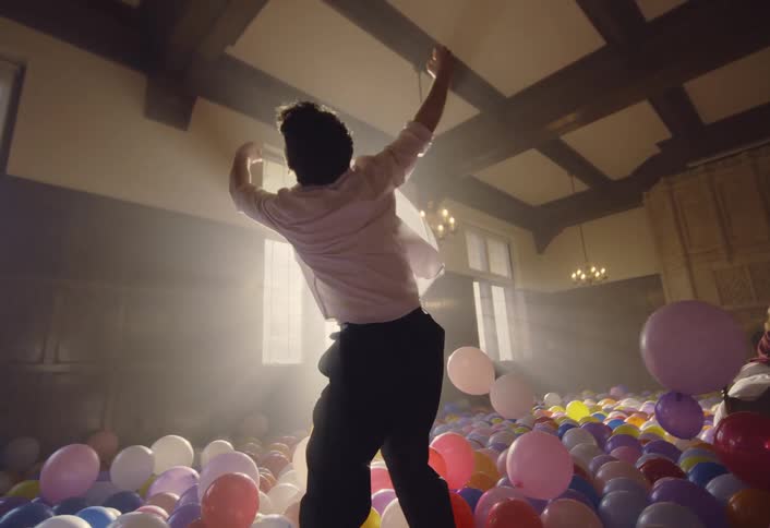 A man dances intensely among colorful balloons in a sunlit hall while a woman sits on a bed in the background