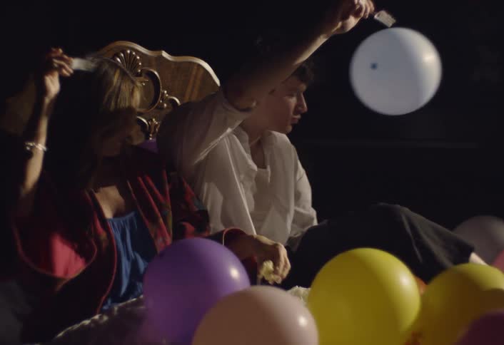 A man and a woman sit on a bed in a dark room while eating cake and dancing among many colorful balloons