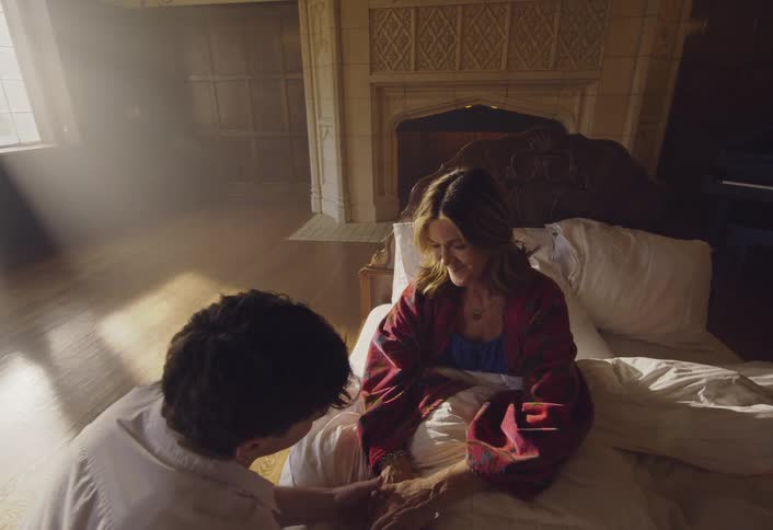 A young man in a white shirt walks across a large room and kneels by a bed to hold the hands of a woman who is sitting up and smiling at him