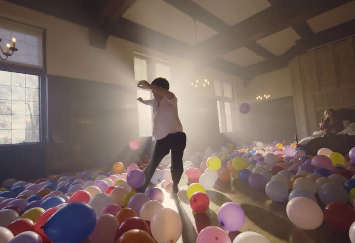 A young man performs a contemporary dance in a large hall filled with scattered balloons and volumetric lighting