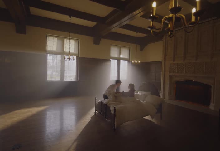 A young man in a black jacket enters a room and kneels beside an elderly woman lying in a bed who has medical tubes in her nose