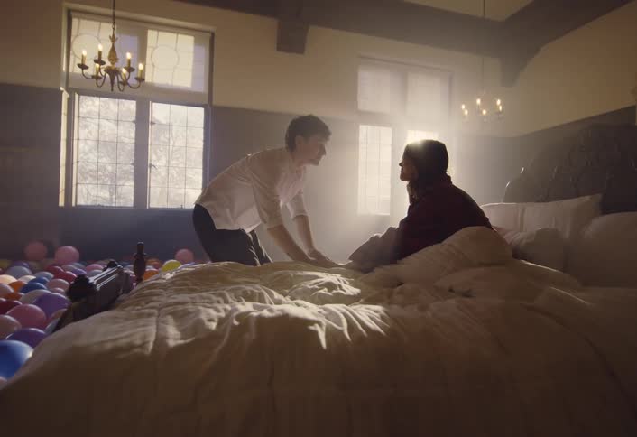 Man holds the hands of a woman sitting up in bed before dancing through the balloons covering the bedroom