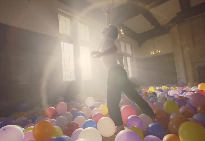A man dances energetically in a sunlit room filled with colorful balloons while another person sits on a bed in the background