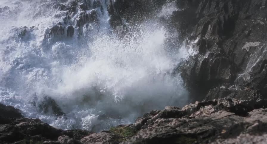 A close view shows powerful ocean waves crashing against dark jagged rocks and splashing white foam into the air