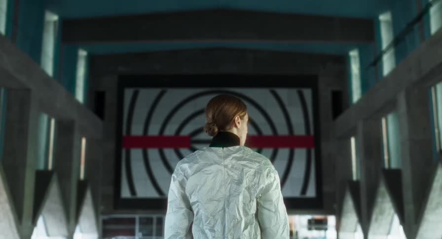 A person in a white jacket stands facing a large screen with bold circular patterns and a red horizontal line in a futuristic interior