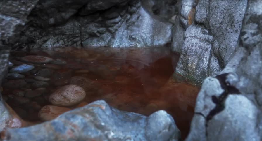 A close view shows a small pool of dark red water tucked within gray jagged rocks where small ripples form on the surface