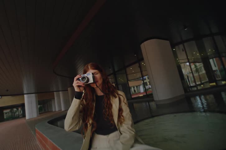 A woman with long hair sits near a curved fountain outside a modern building while holding a camera up to take a photo