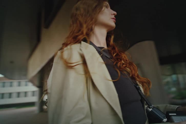 A woman with long ginger hair wearing a beige blazer walks through a modern urban plaza with large concrete pillars while looking around as the camera follows her from a low angle during the day