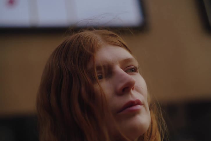 A redheaded woman looks toward the sky with a thoughtful expression while wind blows through her hair against a blurred urban background as she reaches up to tuck a strand behind her ear during a quiet moment outdoors