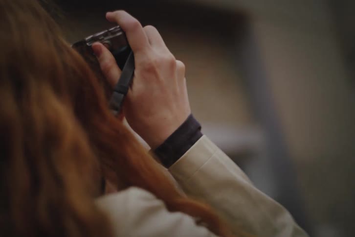 A close view shows a woman with long red hair as she holds a camera to her eye and takes photos of the city around her