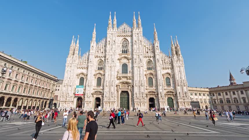 Crowds of people gather in a large city square in front of an ornate white cathedral with towering spires under a clear blue sky