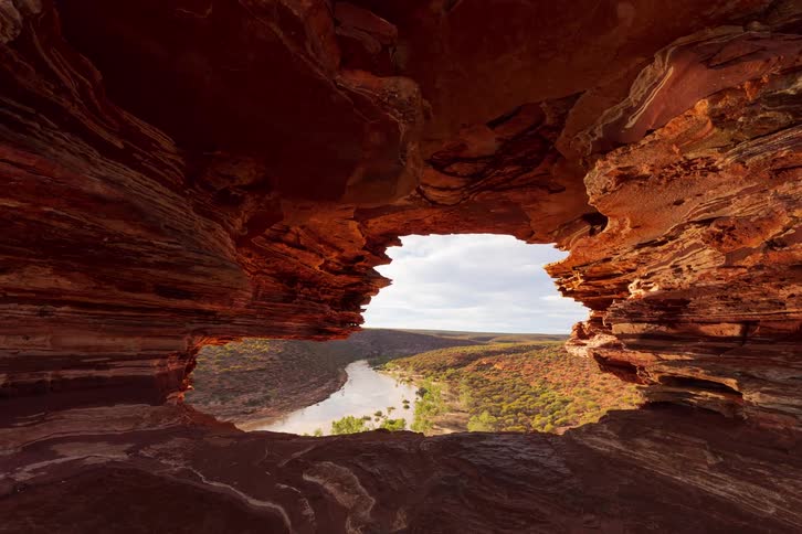 A person looks through a jagged red rock archway toward a winding river that cuts through a vast green valley under a cloudy sky