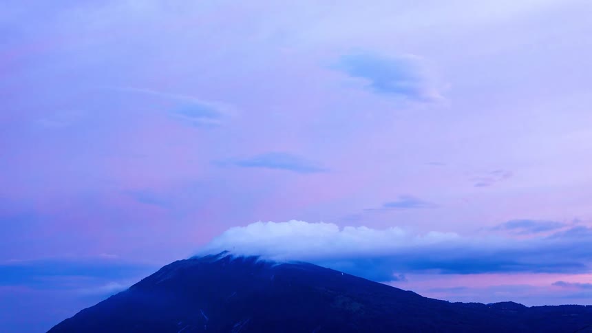 Soft pastel clouds drift above a dark mountain peak as a low band of clouds wraps around its summit