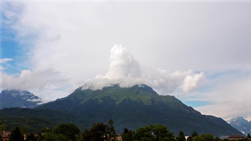 White clouds swirl and dissipate around the emerald green peak of a massive mountain while a small village and thick line of trees sit quietly in the valley under a bright sky