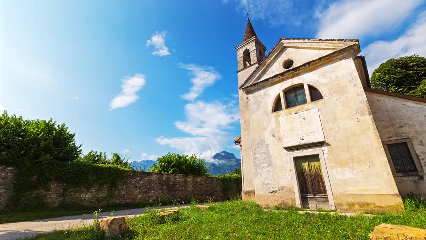 A low view shows an old stone church in a field of grass and flowers with a stone wall and mountains under a bright blue sky