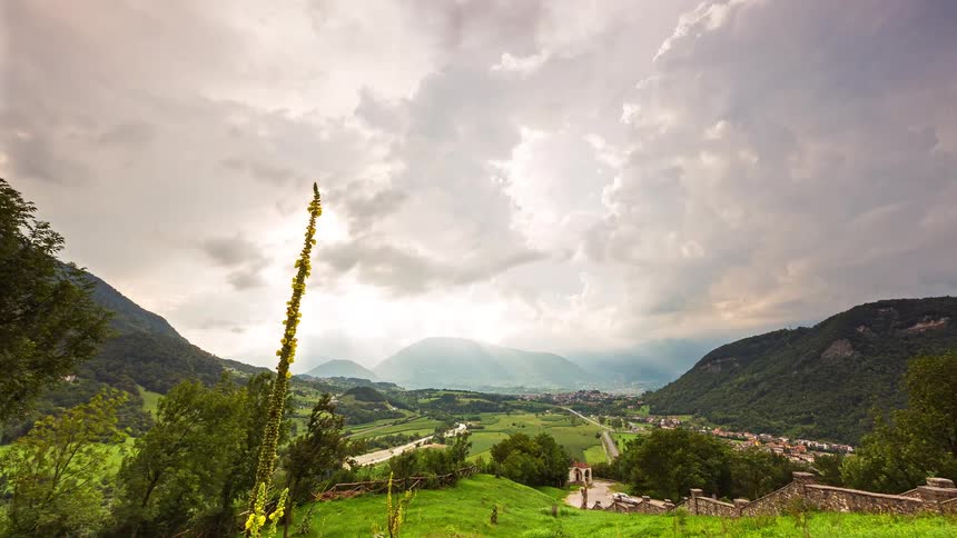 Sunlight breaks through clouds over a lush green valley surrounded by mountains and small rural villages