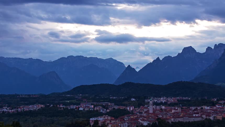 A town with redroofed buildings lies beneath dark clouds with layered mountain peaks rising in the distance