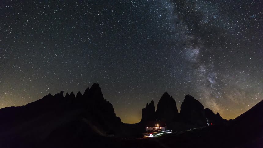 The milky way stretches across a starfilled night sky above dark mountain peaks and a small illuminated building below