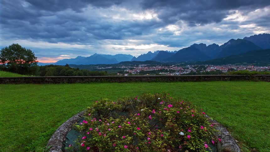 A view from a stone garden with pink flowers shows a wide valley and a city as the sun sets and the sky turns from blue to dark while city lights begin to shine