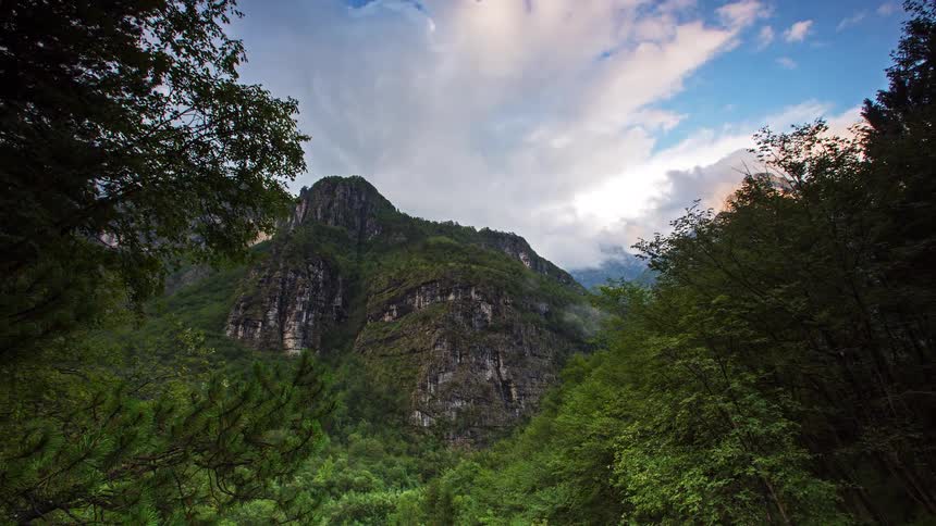 A wide view shows a steep mountain with rocky cliffs rising above a dense green forest under a blue sky with white clouds