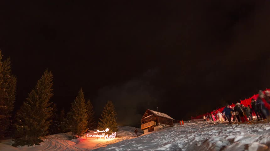 Bright fireworks explode in the night sky over a snow covered mountain with a small wooden cabin and people watching from a ski slope
