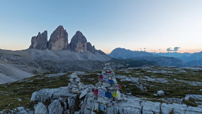 The sun sets behind mountain peaks casting a golden glow over the rocky plateau where small stone cairns decorated with colorful prayer flags stand in the foreground